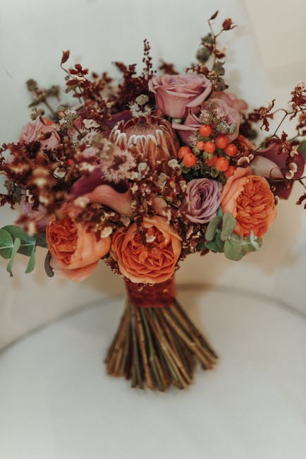 A beautifully arranged bouquet of fresh flowers featuring soft pink roses, vibrant orange carnations, and delicate purple lisianthus, complemented by clusters of small orange berries and sprigs of mut