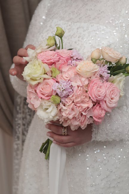 A person wearing a white, sequined wedding dress holds a round bouquet of fresh flowers, featuring soft pink and white roses, pale green buds, and delicate pastel purple blossoms. The bouquet is tight