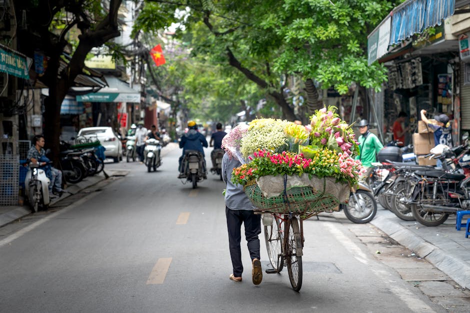 A person pushing a bicycle loaded with a large, vibrant floral arrangement featuring pink, white, and green flowers, including hydrangeas, roses, and foliage. The arrangement is wrapped in rustic, nat