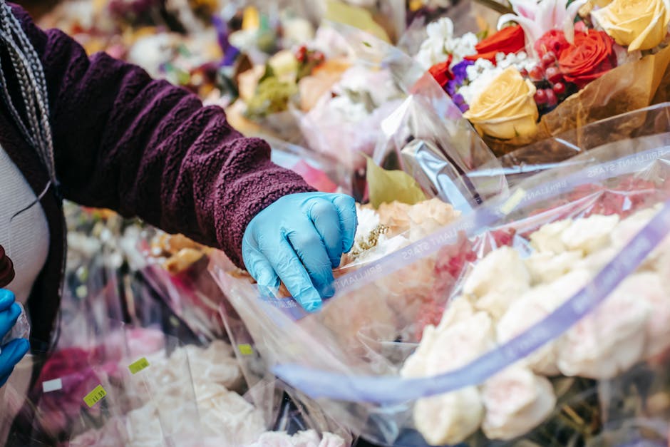 A person wearing a dark purple textured jacket and bright blue gloves is seen handling a clear plastic bag filled with fresh, multicoloured flowers, including white roses, yellow roses, and purple blo