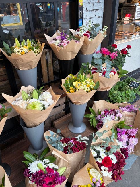 A display of multiple floral arrangements outside a flower shop, featuring a variety of fresh flowers including white lilies, yellow daisies, pink carnations, purple chrysanthemums, and white roses, a