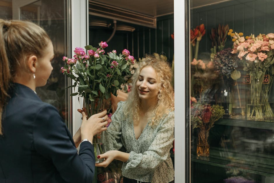 A florist with her brown hair tied back, dressed in a dark blazer, is handing a tall glass vase filled with fresh pink roses to a smiling blonde woman with curly hair, who is wearing a light patterned