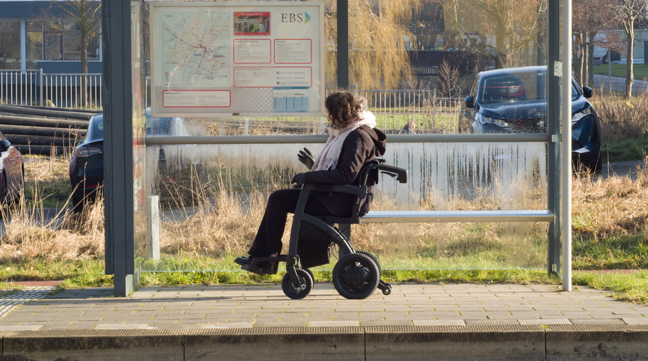 A woman with curly hair wearing a dark coat, pink scarf, and black gloves sits in a wheelchair at a bus stop, facing a large transparent shelter with an information board in the background. The shelte
