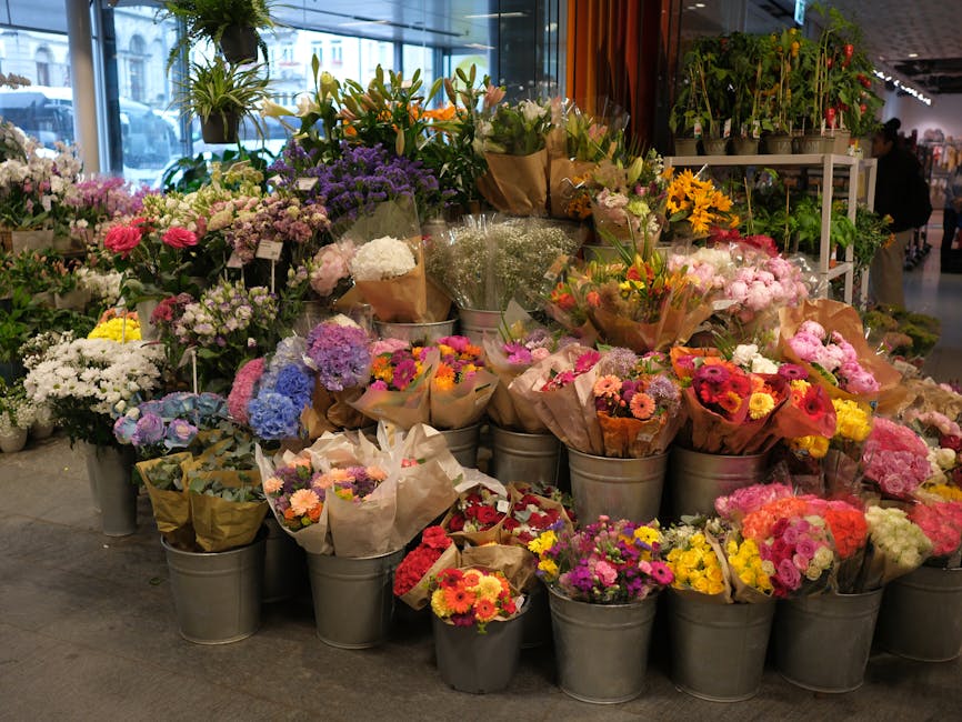 A vibrant display of fresh flowers arranged in galvanized metal buckets and wrapped in brown craft paper, featuring a variety of blooms including pink roses, purple hydrangeas, yellow daisies, white c