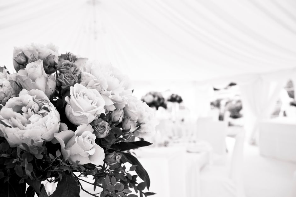 A close-up view of a floral arrangement featuring large, rounded white peonies and pale pink roses, accented with dark green leaves. The bouquet is positioned on a table in an elegant, softly lit sett
