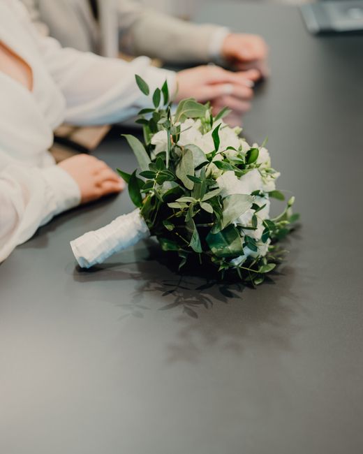 A bouquet of fresh white roses and green foliage, including eucalyptus leaves, lies on a dark grey table surface. The flowers are arranged tightly with visible petal textures and sturdy stems, wrapped