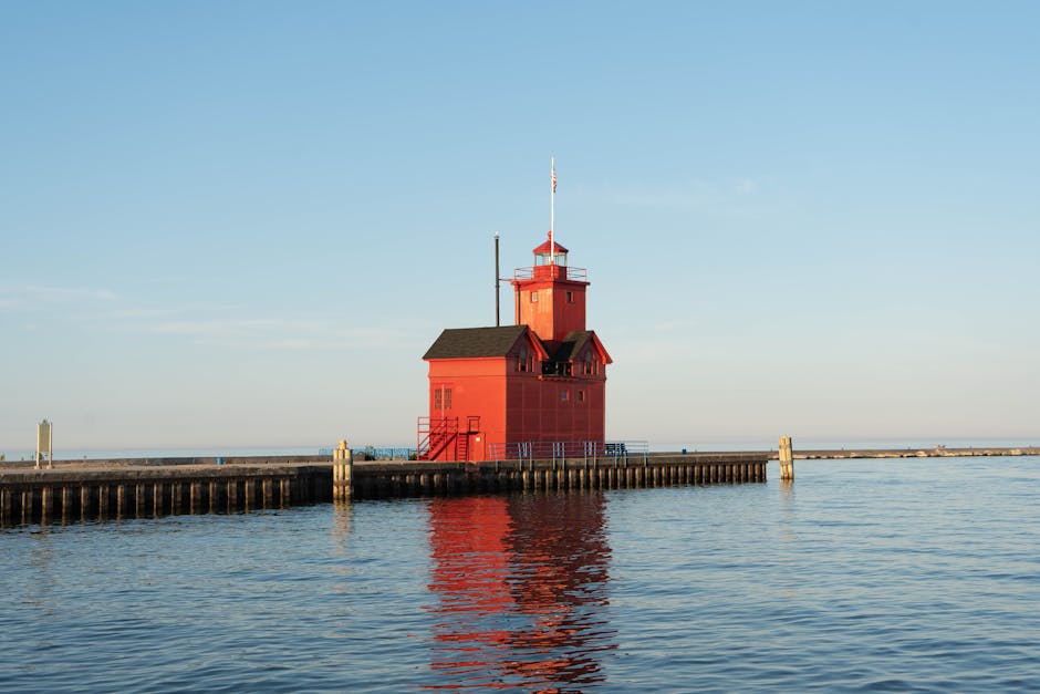 A bright red lighthouse situated on a wooden pier extending into calm water, with a black roof, small windows, and a flagpole on top. The lighthouse is reflected in the water below, and the sky above 